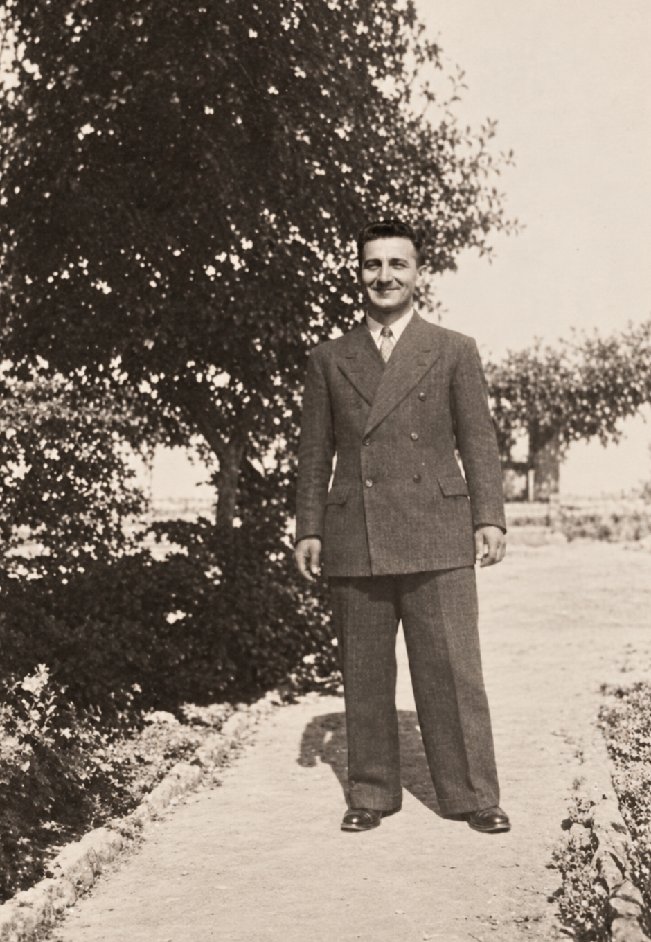 Un uomo in abito posa lungo un vialetto alberato.