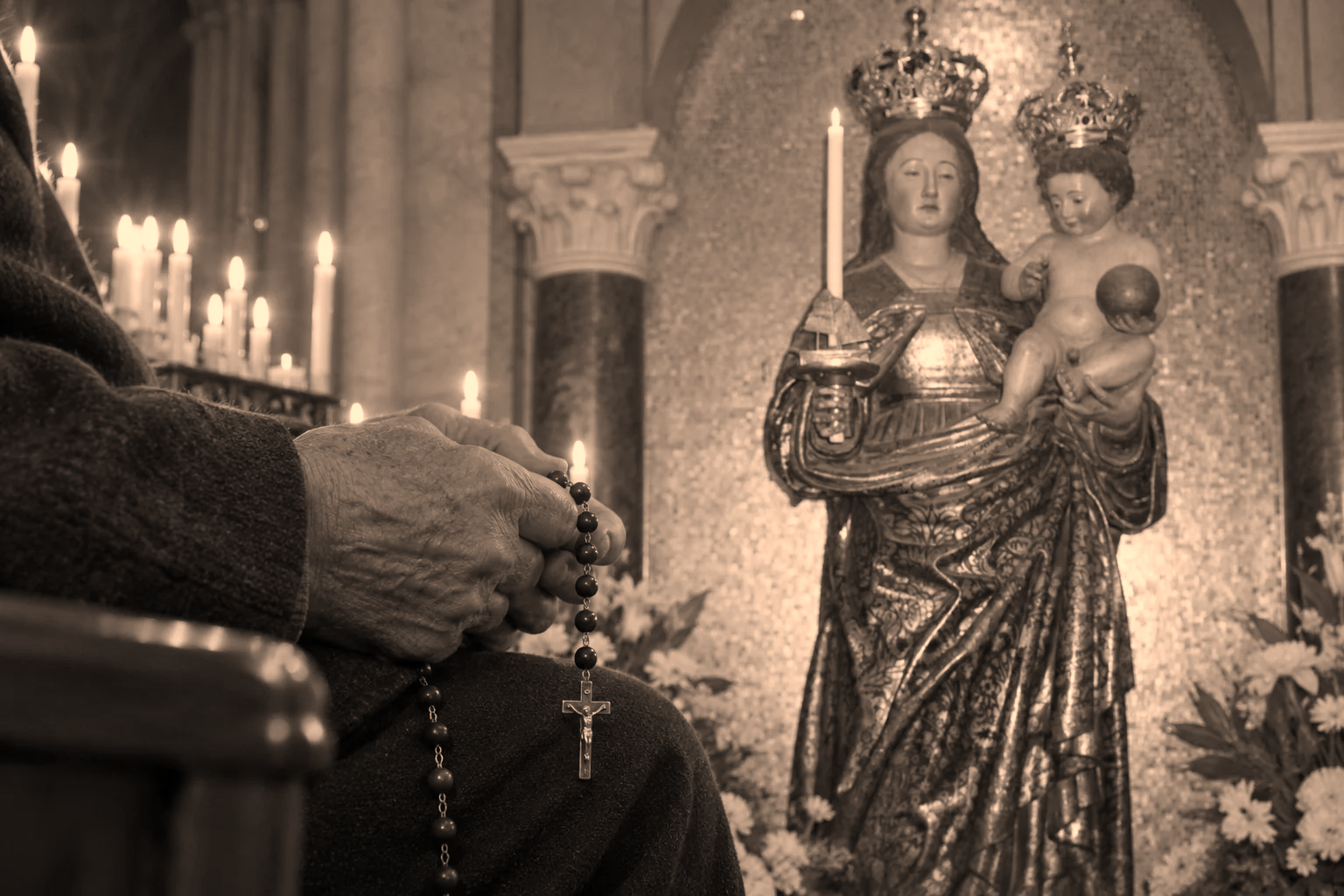 Mani raccolte su un rosario, con candele e una statua della Madonna con il Bambino sullo sfondo.