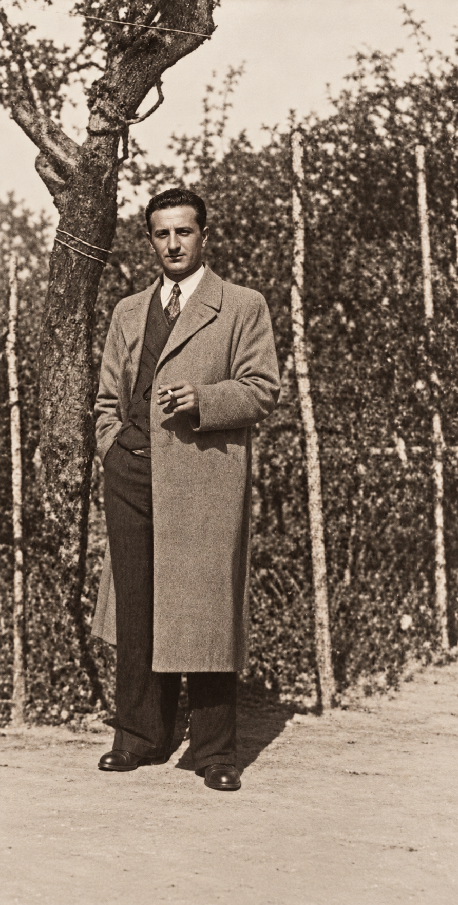 Un uomo in cappotto posa lungo un vialetto vicino a una rete e agli alberi.